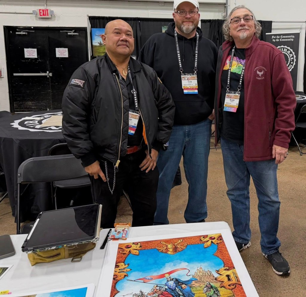 Three men standing together at a convention booth, posing for a photo. They are smiling and wearing casual attire. A colorful piece of artwork is displayed on a table in front of them.