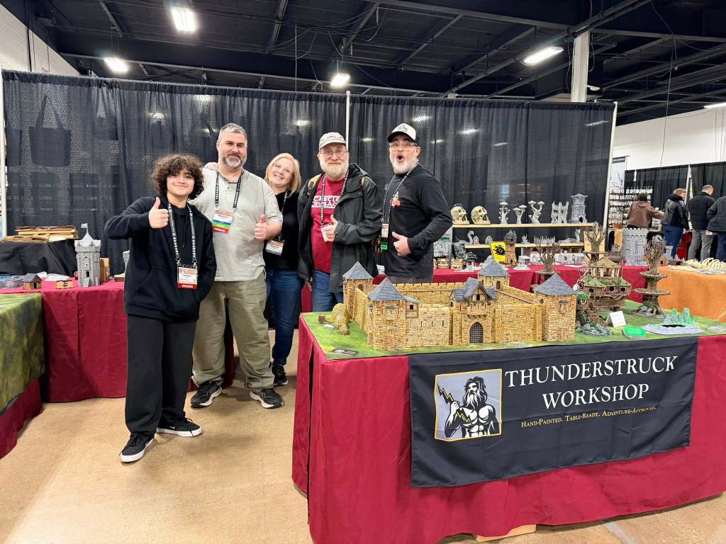 A group of five people posing in front of a vendor booth at a convention, showcasing detailed miniature models and a banner for Thunderstruck Workshop.