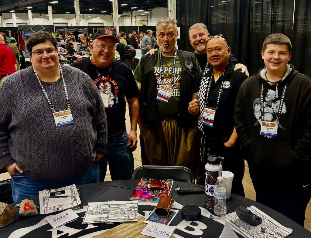 A group of six people poses together at a convention table, smiling and interacting. They are dressed in casual attire and wearing name badges. Various items are visible on the table, including game sheets and a water bottle.