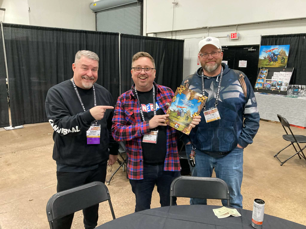 Three men standing together at an event, one holding a board game while smiling. The backdrop features black curtains, tables, and various promotional materials.