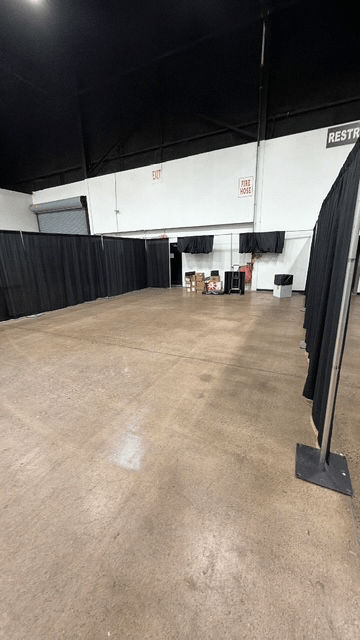 An empty indoor space with a polished concrete floor and black curtains on the sides, featuring a small stack of boxes and a trash can in the background.