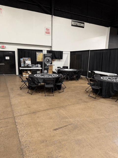 Interior view of an event space featuring several tables with black tablecloths and folding chairs, along with displays and a sign indicating restrooms.