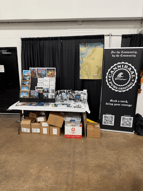A vendor booth showcasing Cannibal Publishing materials at an event. The display includes books, promotional posters, a large banner, and boxes of merchandise, set against a black backdrop.