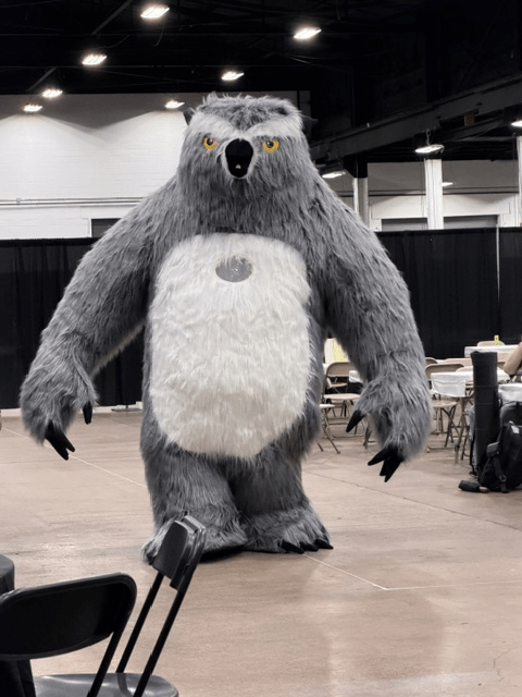 A large gray bear mascot costume walking in a convention hall with tables and chairs in the background.