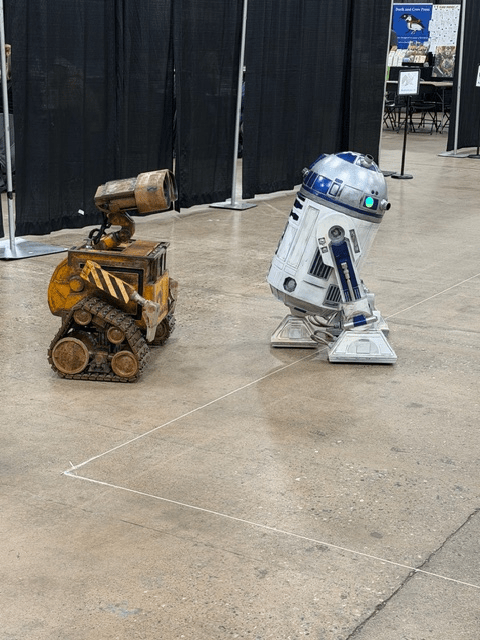 Two robotic characters in a convention setting: a small, rusty, yellow and brown robot resembling Wall-E and a tall, white and blue R2-D2 robot.