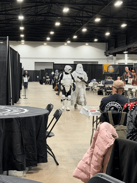 Two costumed characters walk through a convention center, one in a stormtrooper outfit and the other in a white costume, with various tables and attendees visible in the background.