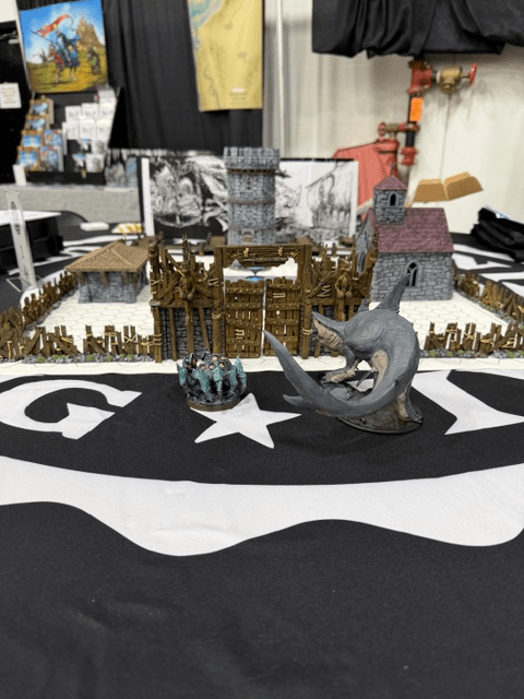 A tabletop gaming scene featuring a detailed miniature fortress with wooden walls, towers, and a church, surrounded by a group of miniature figures and a large creature figure. The background includes a black and white star-patterned fabric.