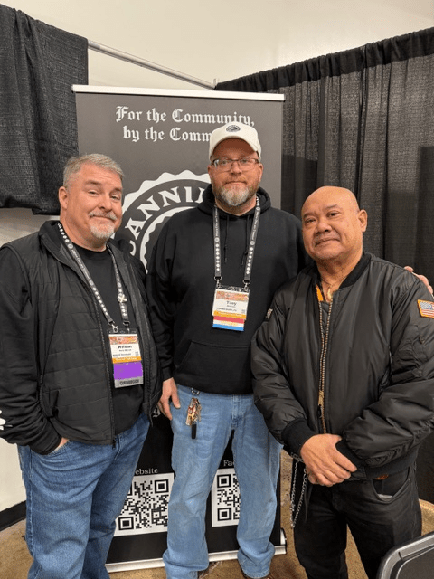 Three men pose for a photo at an event, standing in front of a display banner that reads 'For the Community, by the Community'.