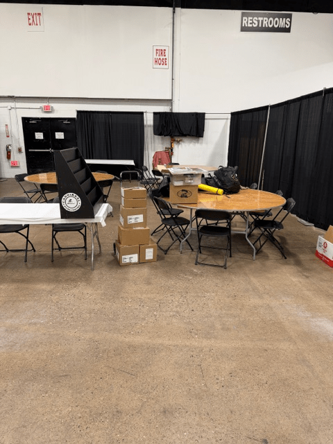 An empty event space featuring several tables and chairs, with stacked boxes nearby and black curtains in the background. Signs for restrooms and exits are visible.