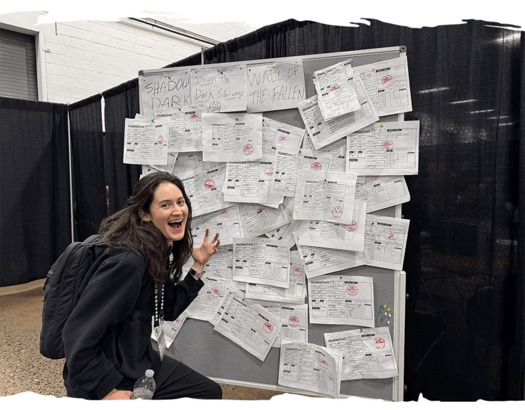 A smiling woman in black clothing with a backpack stands next to a bulletin board covered in various papers and forms, labeled 'Wall of the Fallen'.