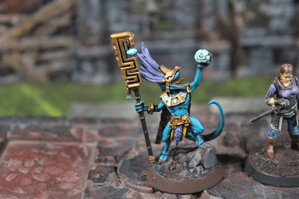 A colorful, detailed miniature of a character resembling a humanoid creature with blue skin and a headdress, holding a staff and a glowing orb, posed on a rocky base next to another humanoid figure in a purple outfit.