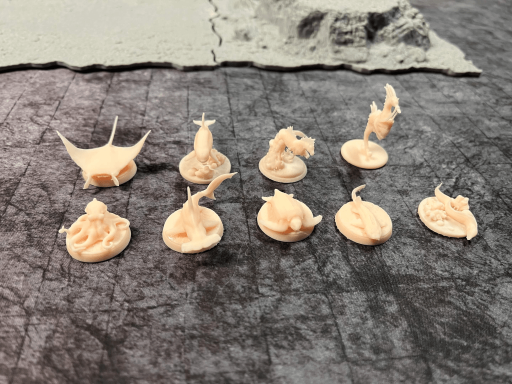 A collection of 10 small, unpainted fantasy-themed miniatures featuring various sea creatures like octopuses, mermaids, and other aquatic figures, arranged on a textured surface.