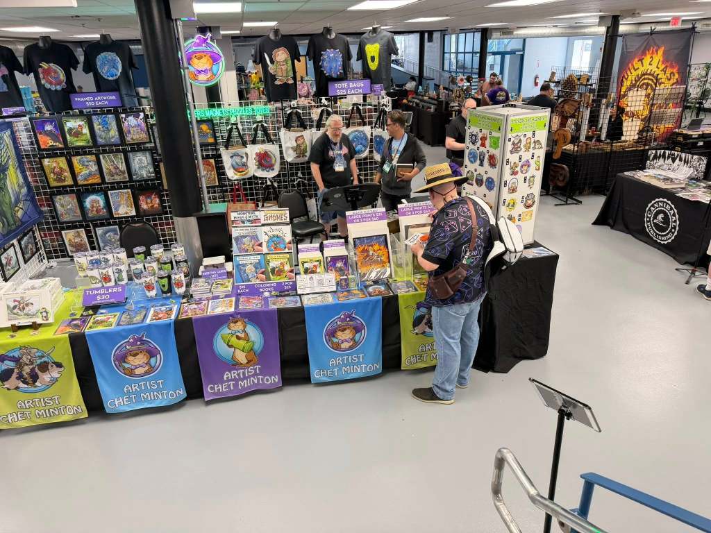 A vibrant artist booth showcasing merchandise such as artwork, tumblers, and coloring books, with colorful banners displaying the artist's name. Attendees browse the products while various artistic items are displayed on nearby tables.