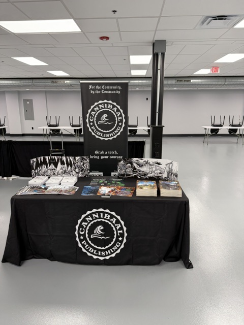 Table displaying materials from Cannibal Publishing, with a black tablecloth featuring the logo, a banner in the background, and various magazines and brochures arranged neatly.