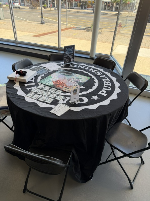 A round table covered with a black tablecloth featuring a logo, surrounded by several empty chairs. On the table, there are various printed materials, including a colorful map and flyers, along with a few loose papers.
