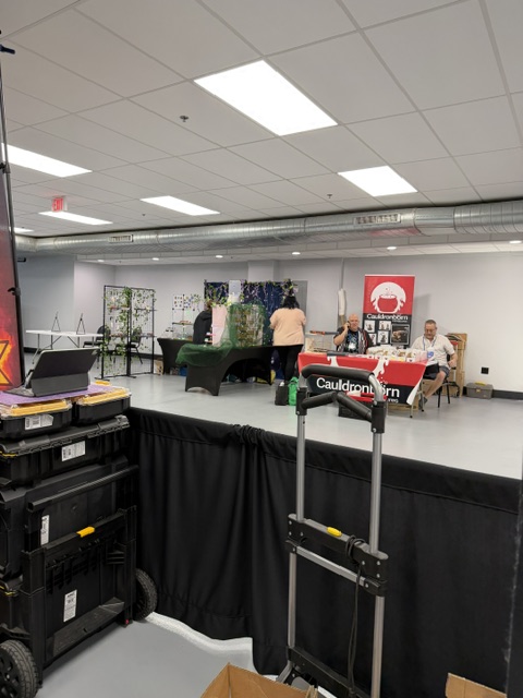A view of an indoor event space featuring vendors and displays. In the foreground, there are stacked boxes and a cart, while in the background, several people are attending to tables with plants and a red Cauldron Corn banner.