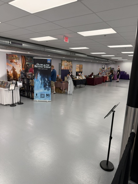 A spacious convention hall with vendor tables showcasing various games and merchandise, featuring a promotional banner for 'Glory in the Round Table'.