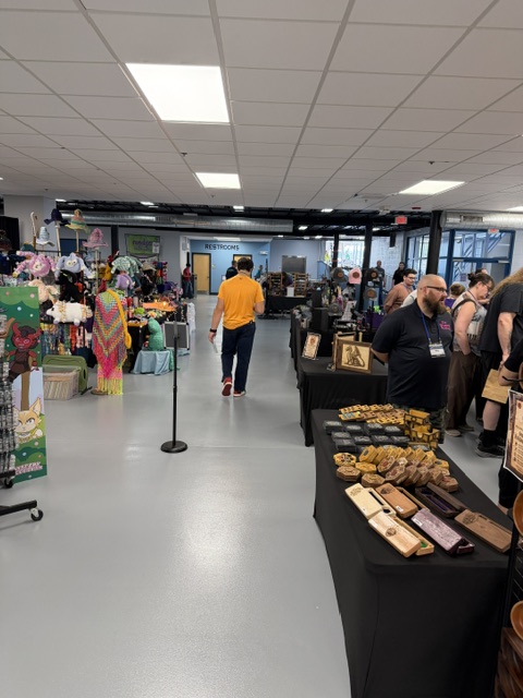 A view of a vibrant indoor market with various vendor booths showcasing colorful merchandise and crafts. People are browsing the stalls, with a mixture of handmade items and art on display. Restrooms can be seen in the background.
