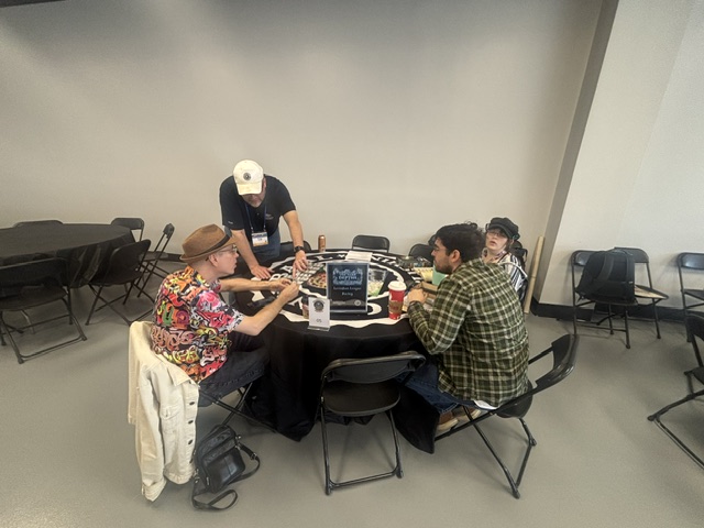 Four people gathered around a table discussing and engaging in an activity, with some food and drinks present.