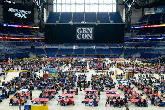 A wide view of a large convention center filled with people attending GEN CON, featuring numerous tables and booths set up for various activities and games.