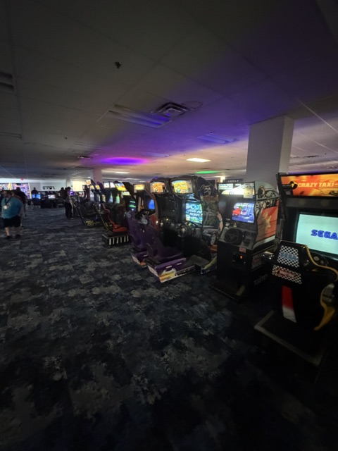 A row of arcade machines in a dimly lit gaming area, with colorful screens and various game titles visible, surrounded by carpeted flooring and soft lighting.