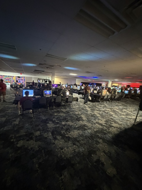 A dimly lit gaming room with numerous computer stations, each with monitors displaying various screens. Several individuals are seated at the desks, engaged in gaming activities. The atmosphere is vibrant with colored lights in the background.