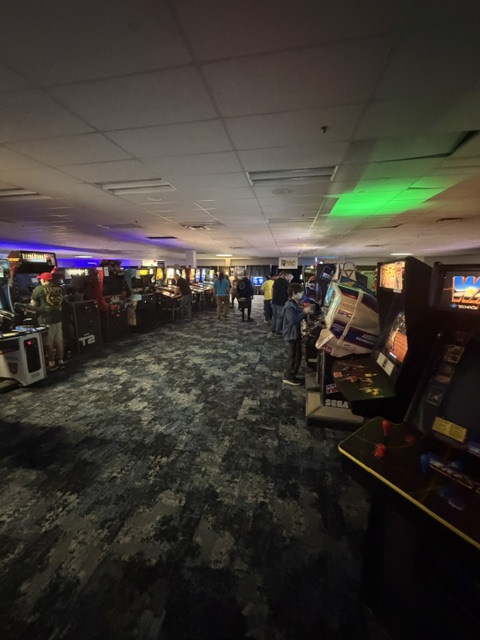 Interior of an arcade with multiple rows of classic arcade machines, dim lighting, and a few people interacting with the games.