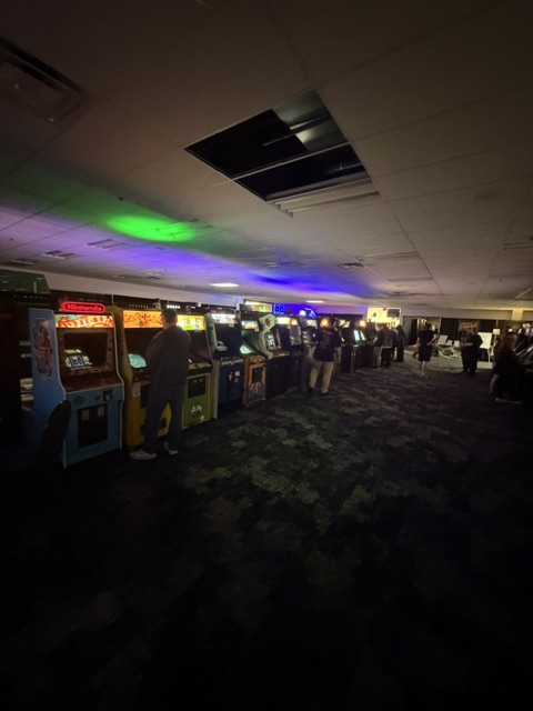 A dimly lit arcade featuring a row of vintage arcade game machines with colorful screens and illuminated controls. People are engaged with the games, creating a nostalgic atmosphere.