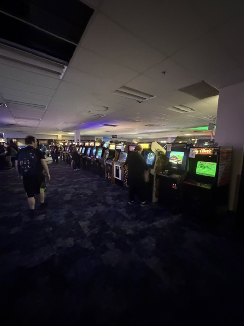 A dimly lit arcade filled with various vintage arcade game machines along the wall, with people playing and walking around.