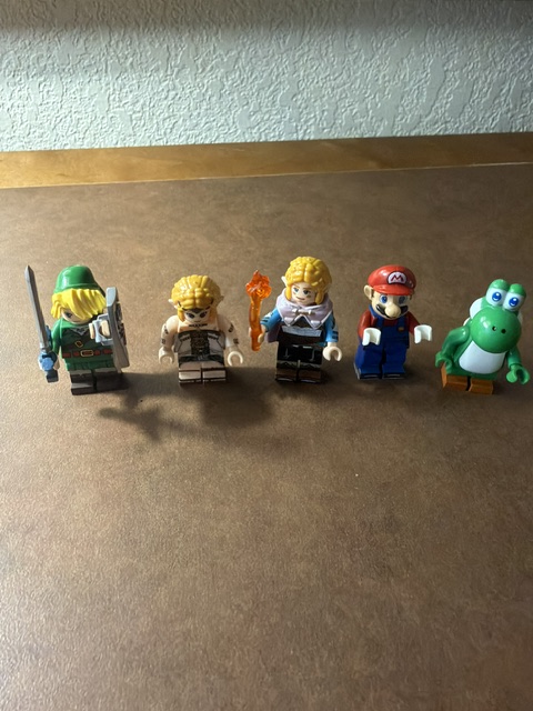 Five LEGO figurines standing in a row on a brown surface. The first figure is a warrior holding a sword and shield, the second is wearing a simple outfit, the third has a fire torch, the fourth is a character in a red hat and overalls, and the fifth is a green dinosaur with big eyes.