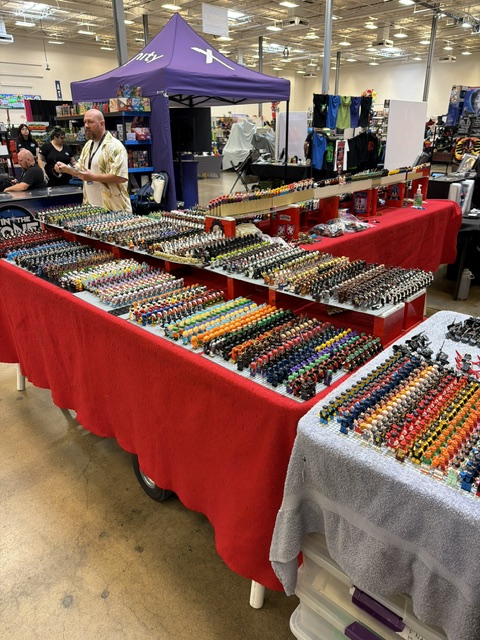A vendor booth at a convention displaying a large collection of colorful miniature figures on tables covered with red tablecloths.