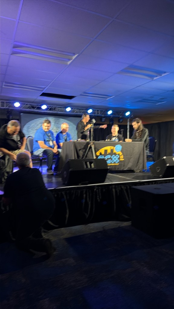 A panel discussion taking place on stage with multiple speakers sitting at a table, under bright stage lights, in a conference setting.