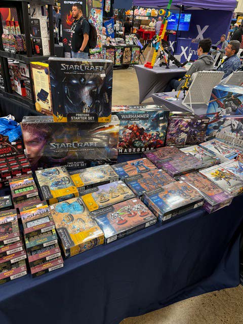 A display of various tabletop gaming products, including 'StarCraft' starter sets and 'Warhammer' games, arranged on a table at a gaming convention.