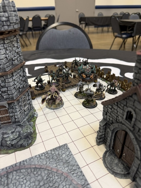 A tabletop game scene featuring miniature figures, including various creatures and structures, set on a grid mat. The setup includes a tower and a fortress entrance.