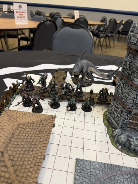 A tabletop gaming scene featuring various painted miniature figures including humanoid characters and a large creature, set against a grid-patterned game mat and a detailed stone tower.