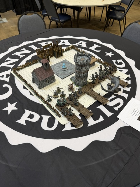 A tabletop gaming setup featuring detailed miniature scenery including a tower, a building, and various figures, displayed on a circular table with a logo of 'Cannibal Publishing'.