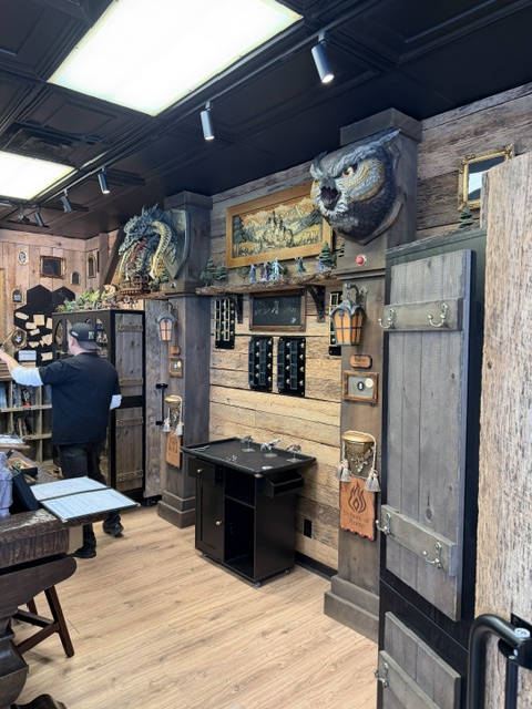 Interior of a rustic-themed shop featuring wooden walls, fantasy decor including a dragon and owl heads, and various handcrafted items displayed. A person in a black shirt is examining a menu at a counter.