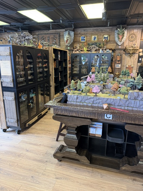 Interior view of a craft store showcasing display cabinets filled with miniature figures, scenic models, and decorative items, with wooden furnishings and a warm ambiance.