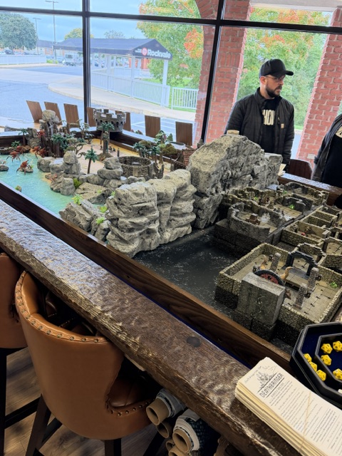 A tabletop gaming setup featuring a detailed landscape with rocky formations, palm trees, and a ruined castle, along with gaming accessories and two individuals in the background.