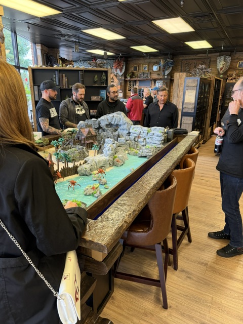 A group of people gathered around a detailed tabletop game setup featuring miniature landscapes and figures in a game store.