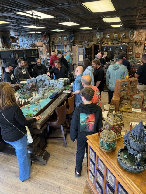 A crowded gaming store interior featuring a long table with a detailed tabletop gaming setup, surrounded by people engaged in conversation and gameplay. Decorative items and shelves line the walls, creating a vibrant gaming atmosphere.