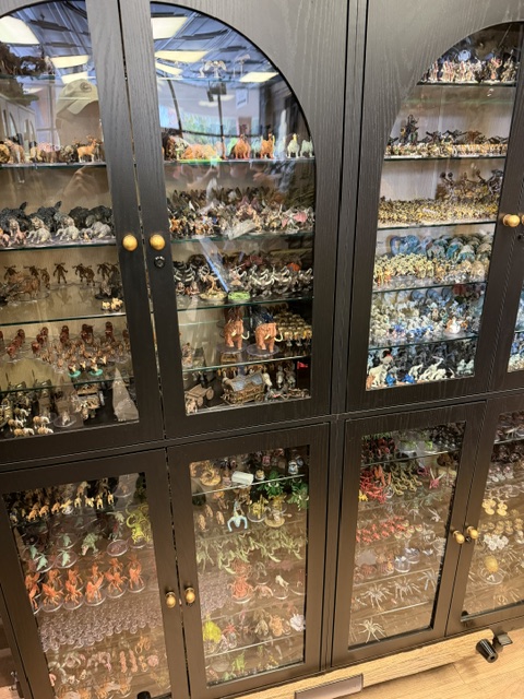 A display cabinet filled with a variety of miniature figures, including fantasy and sci-fi characters, arranged on multiple shelves.