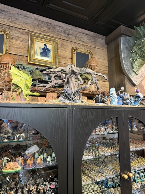 A display case filled with various collectible figurines, including fantasy creatures like dragons and humanoid figures, against a wooden wall with framed artwork and decorative elements.