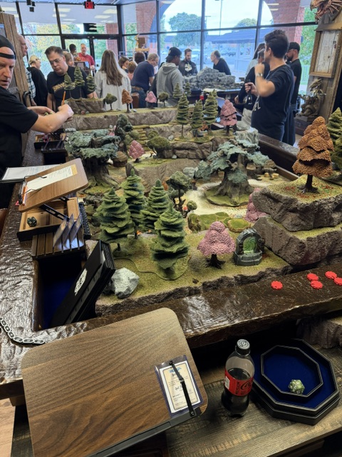 A tabletop gaming scene featuring a detailed landscape with trees, rocks, and miniature structures, surrounded by a crowd of people engaged in gameplay.