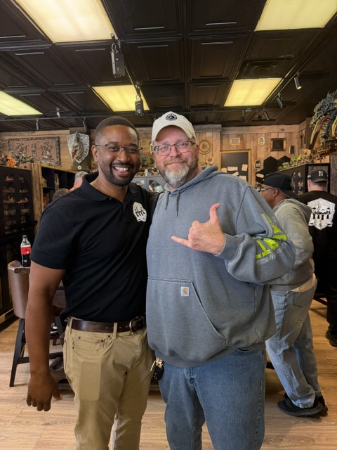 Two men smiling and posing for a photo inside a shop, with a cozy interior filled with various decorations and items in the background.