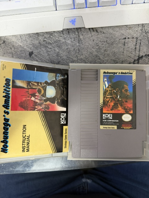 A NES video game cartridge for 'Nobunaga's Ambition' alongside its instruction manual, resting on a tabletop near a keyboard.